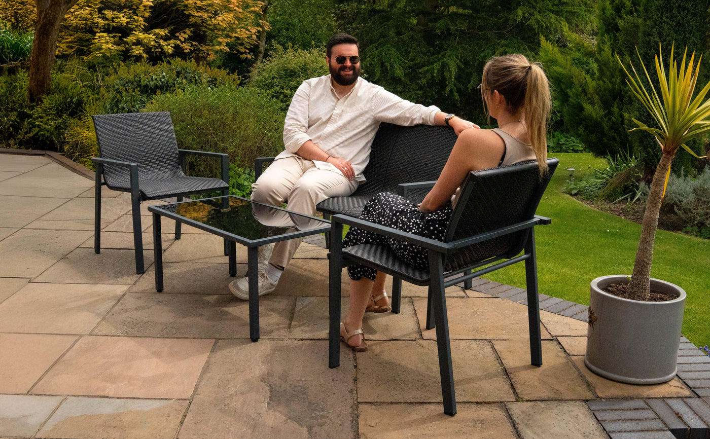 Two people relax on a patio, seated in modern black wicker chairs with a glass table, surrounded by lush greenery and sunlight