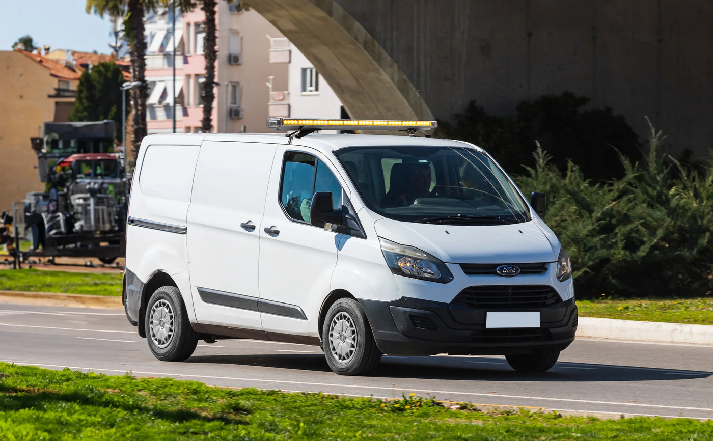 White Ford Transit van with a yellow light bar driving on a sunny street, surrounded by greenery and buildings in the background