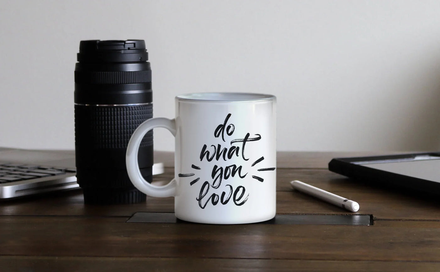 White ceramic mug with "do what you love" in stylish black script, placed on a wooden table next to a camera lens and laptop