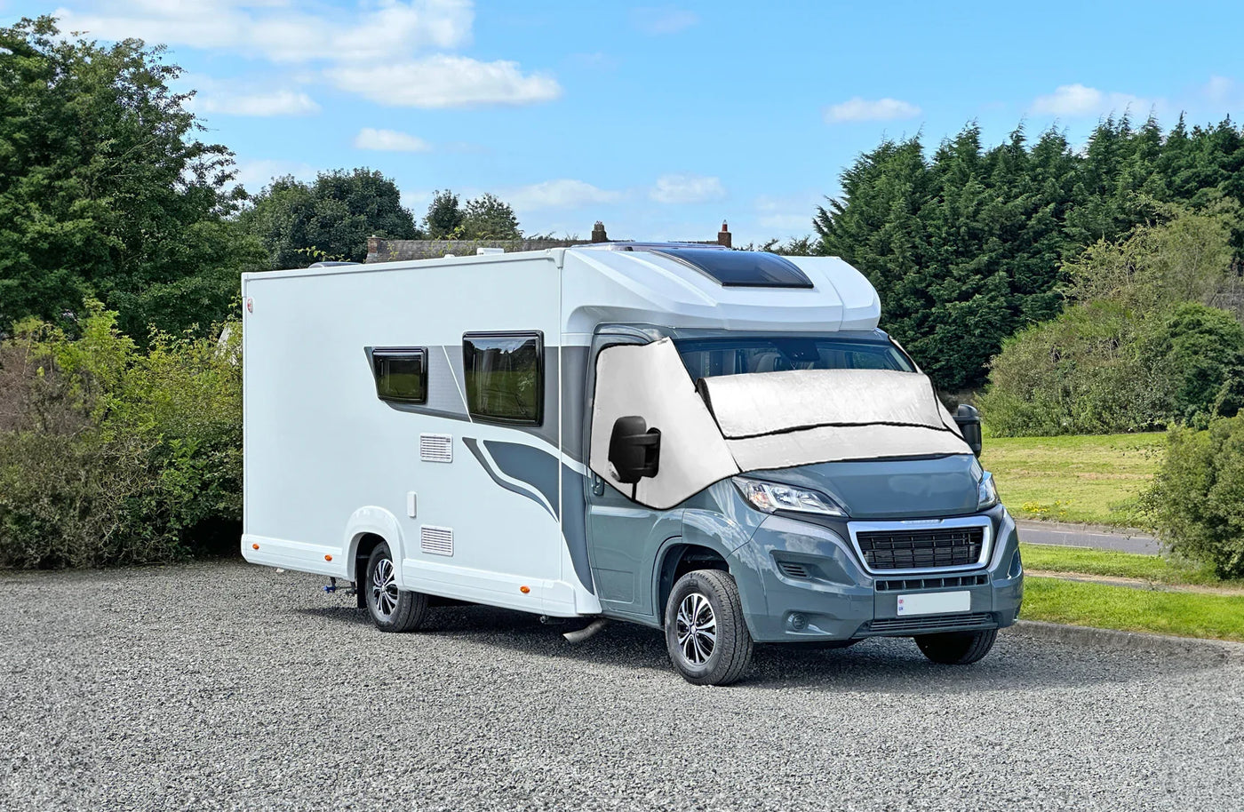 Modern gray and white camper van parked on gravel, featuring large windows, solar panels, and a sleek aerodynamic design against a lush green backdrop