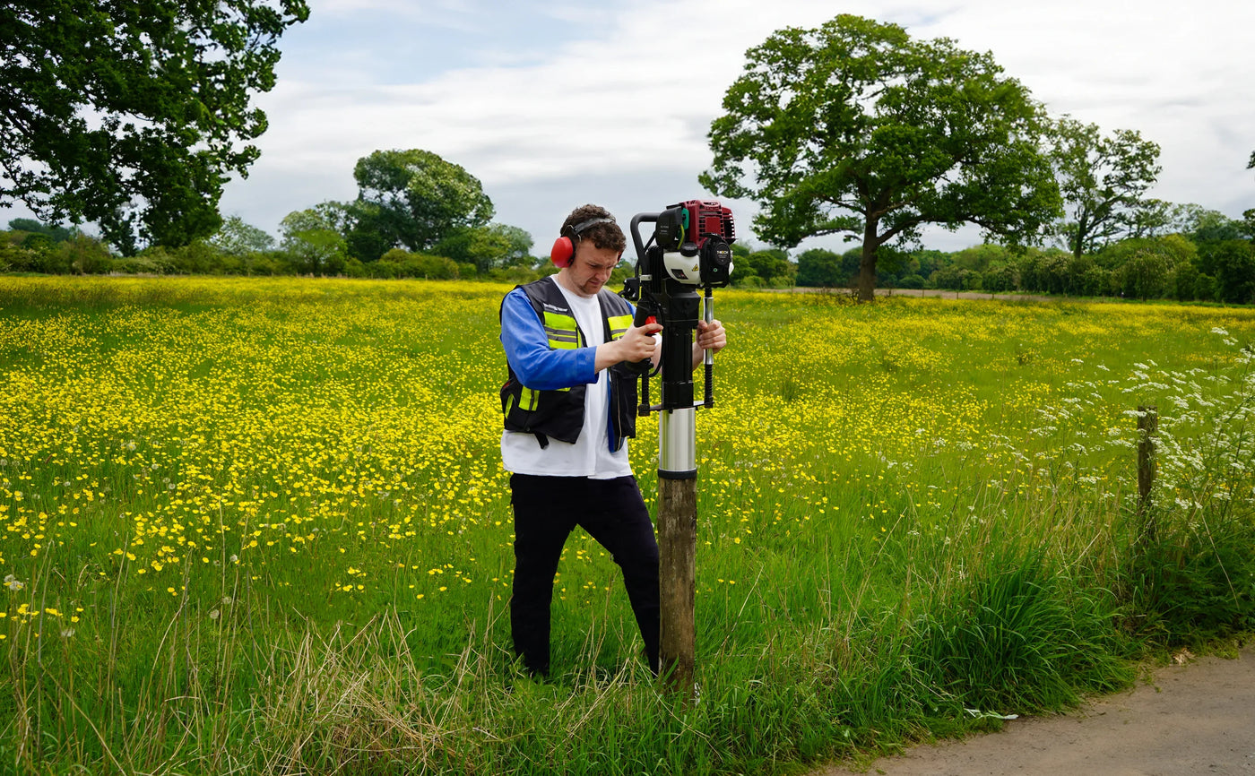 A worker in a safety vest uses a surveying instrument in a lush green field filled with yellow flowers and trees in the background