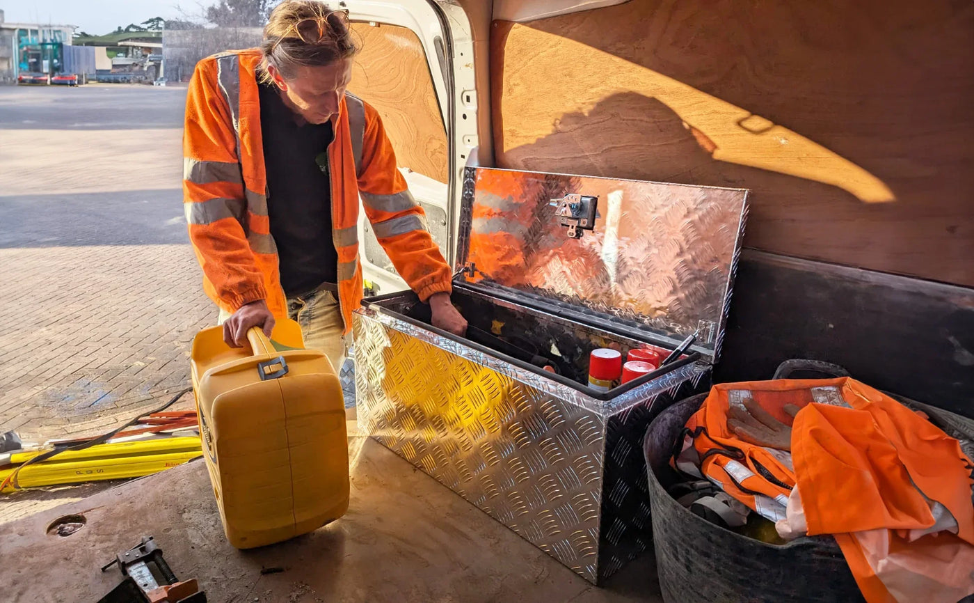 A worker in an orange reflective jacket opens a shiny metal toolbox inside a van, surrounded by tools and safety gear