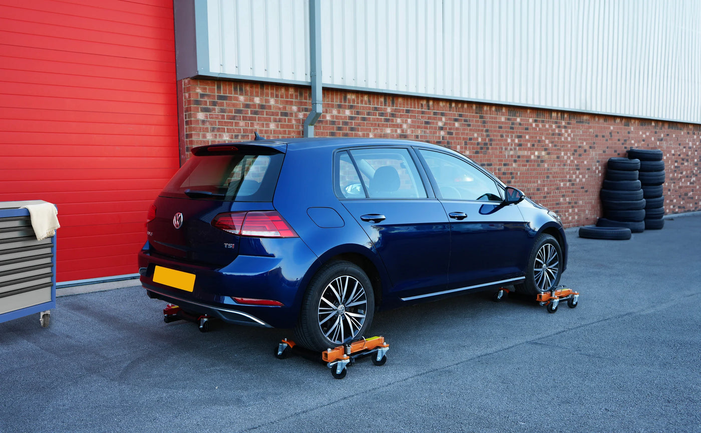 A blue Volkswagen Golf parked on orange wheel dollies outside a brick building, with red garage doors and black tires in the background