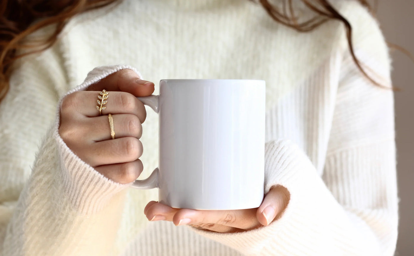 A person in a cozy cream sweater holds a plain white mug, showcasing delicate gold rings on their fingers against a soft background