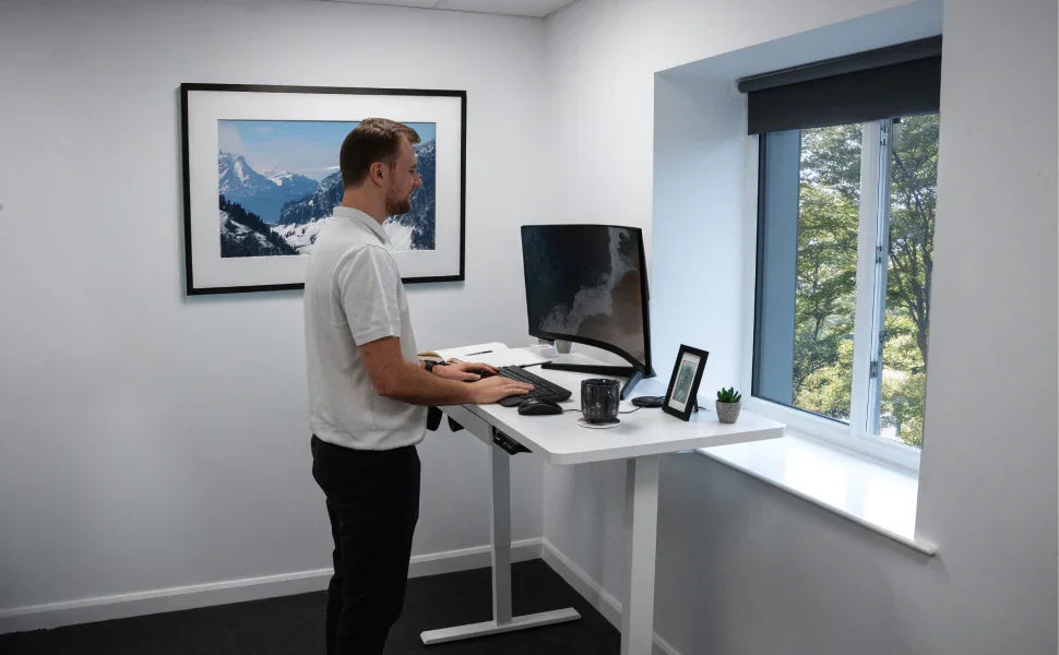 Modern workspace featuring a man at a standing desk, sleek monitor, and potted plant, with a scenic window view and framed mountain photo