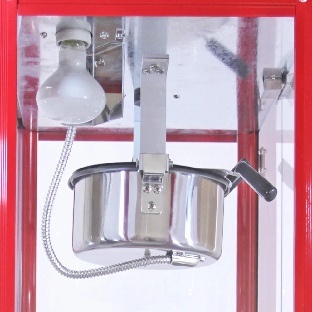Stainless steel kettle with a black handle inside a red popcorn machine, featuring a light bulb above for illumination