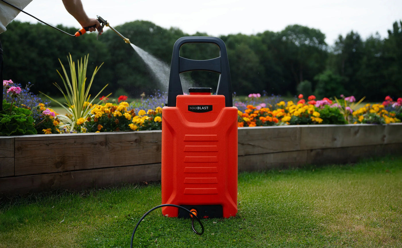 Bright orange electric pressure washer positioned in a lush garden, surrounded by colorful flowers and green grass, with water spraying from a nozzle