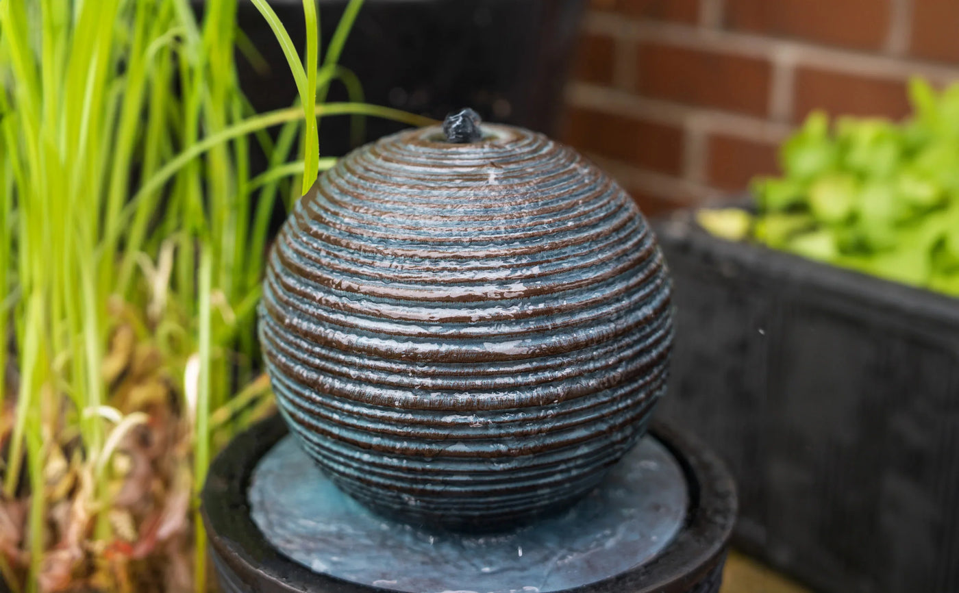 Round, textured water fountain with dark blue and bronze stripes, surrounded by lush green plants in a garden setting