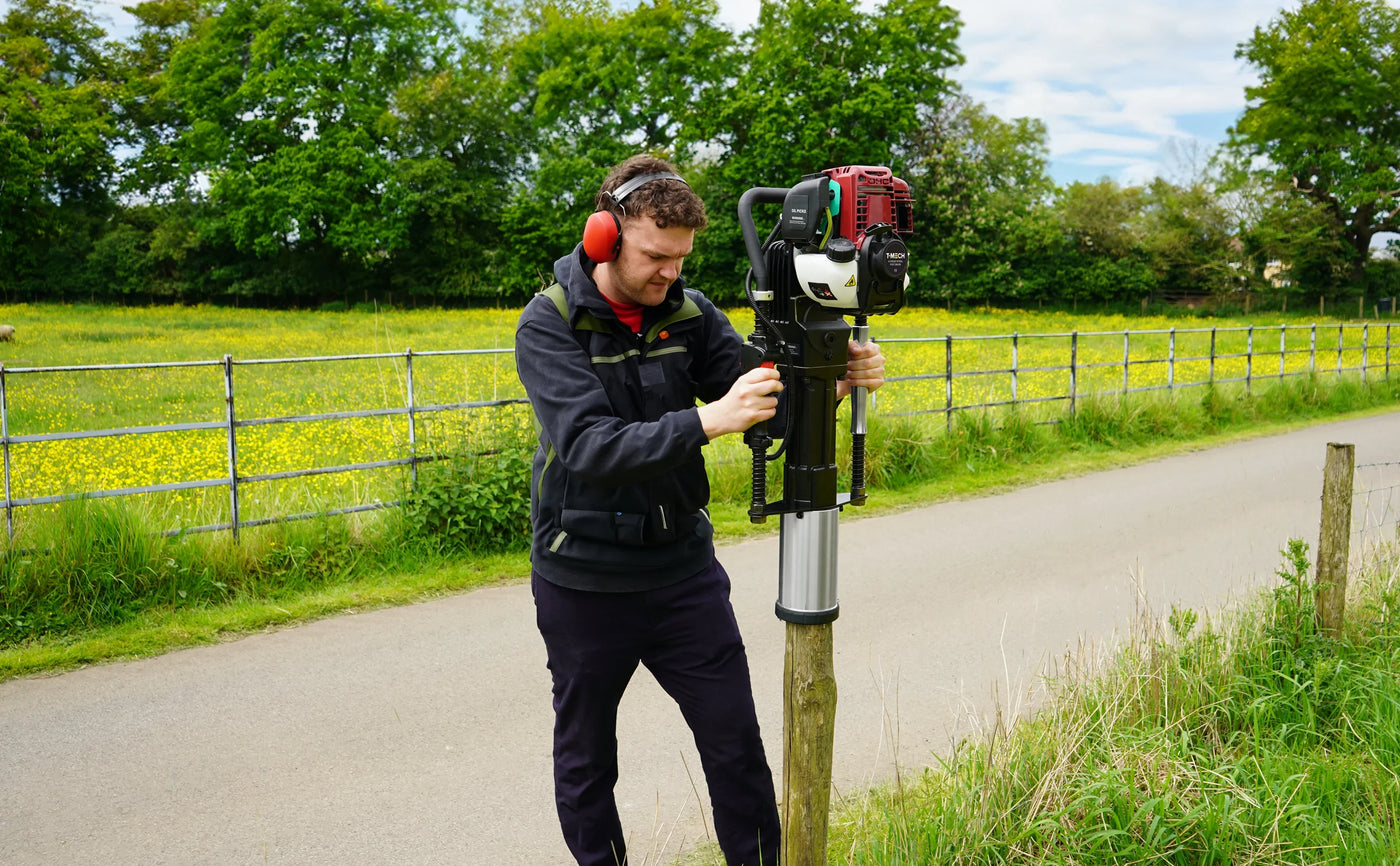 Man using a gas-powered post hole digger on a sunny day, surrounded by green fields and trees, wearing ear protection and a black jacket