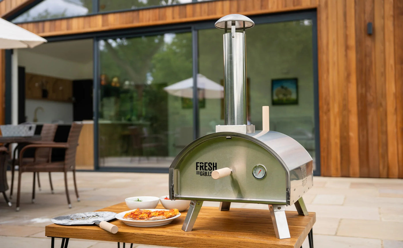 Stainless steel outdoor pizza oven on a wooden table, with fresh pizza and dipping sauces, set against a modern home backdrop