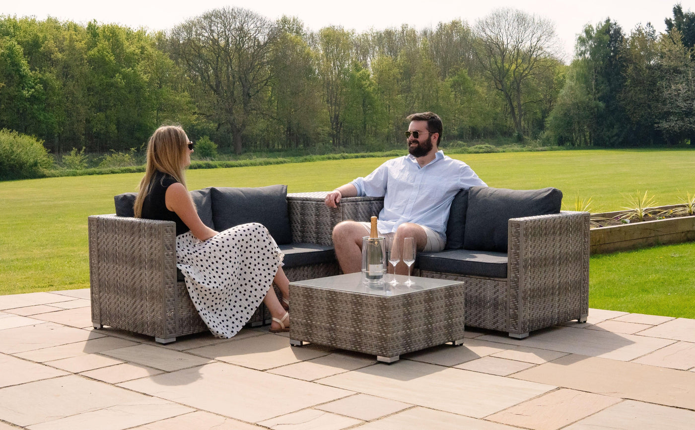 Stylish outdoor seating set with gray wicker chairs and a glass-top table, surrounded by lush greenery and bright sunlight