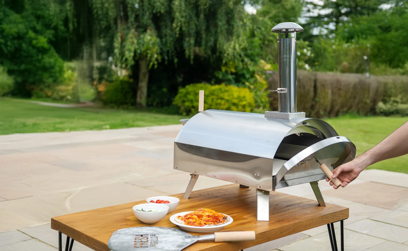 Stainless steel outdoor pizza oven on a wooden table, with fresh pizzas and toppings, set in a lush garden backdrop