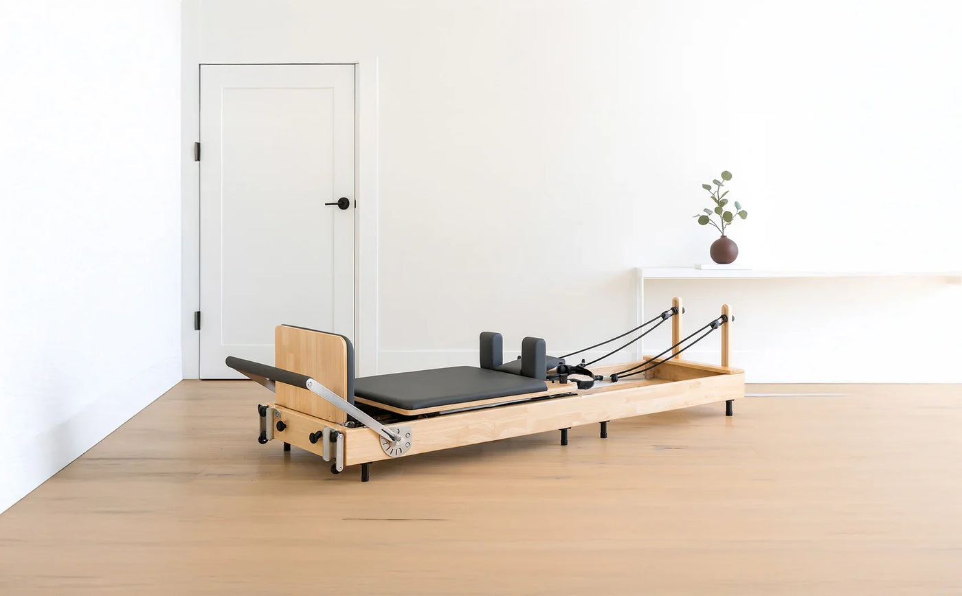 A wooden Pilates reformer with a black padded surface, adjustable straps, and a minimalist design, set in a bright, airy room