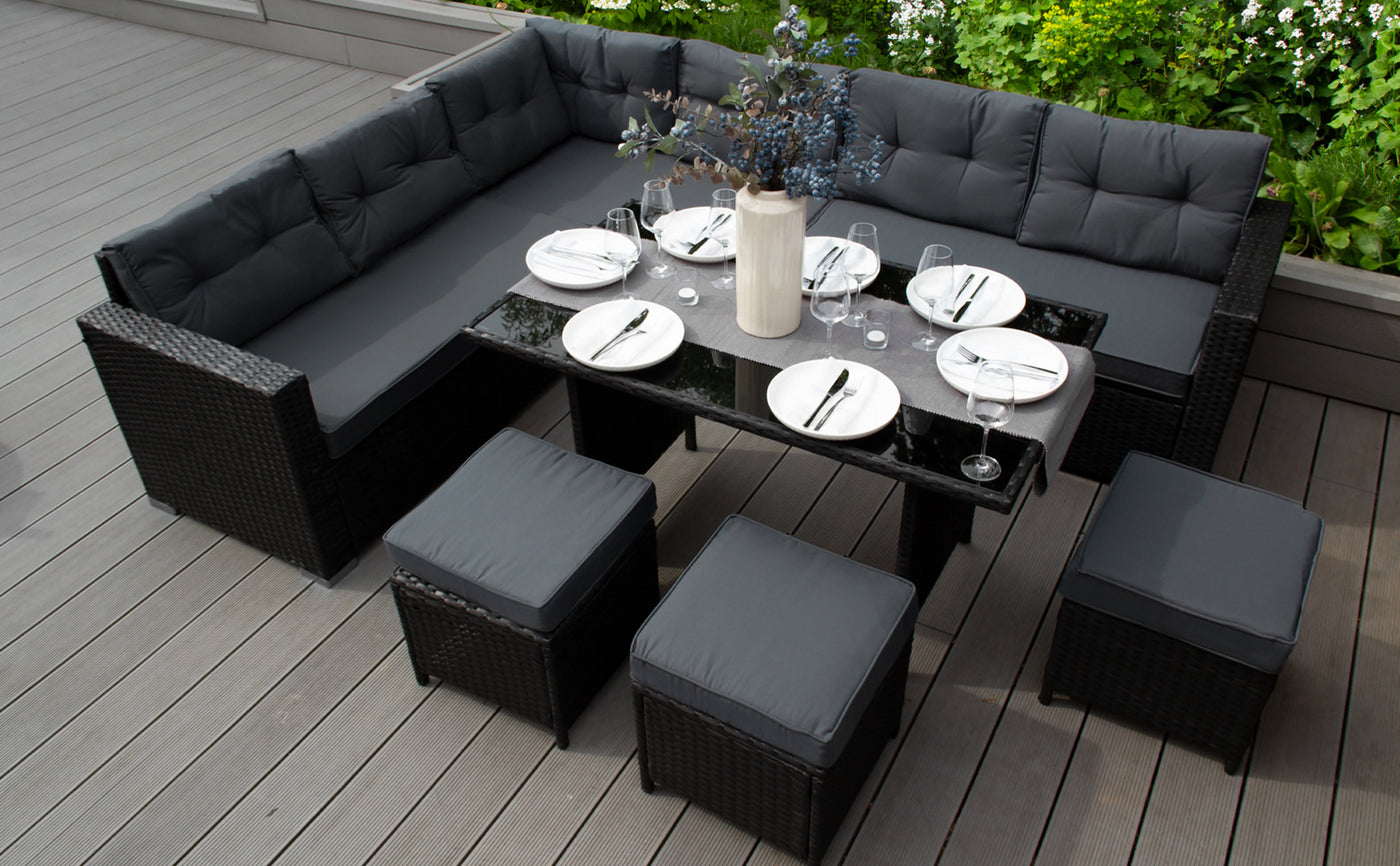 Stylish outdoor dining set featuring a black wicker sectional sofa, glass table, and matching stools, surrounded by greenery