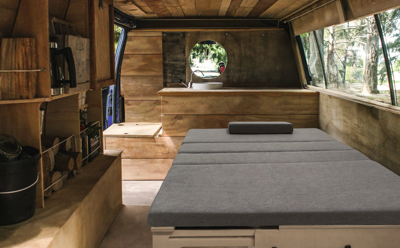 Cozy interior of a camper van featuring a gray mattress, wooden cabinetry, and large windows with natural light streaming in