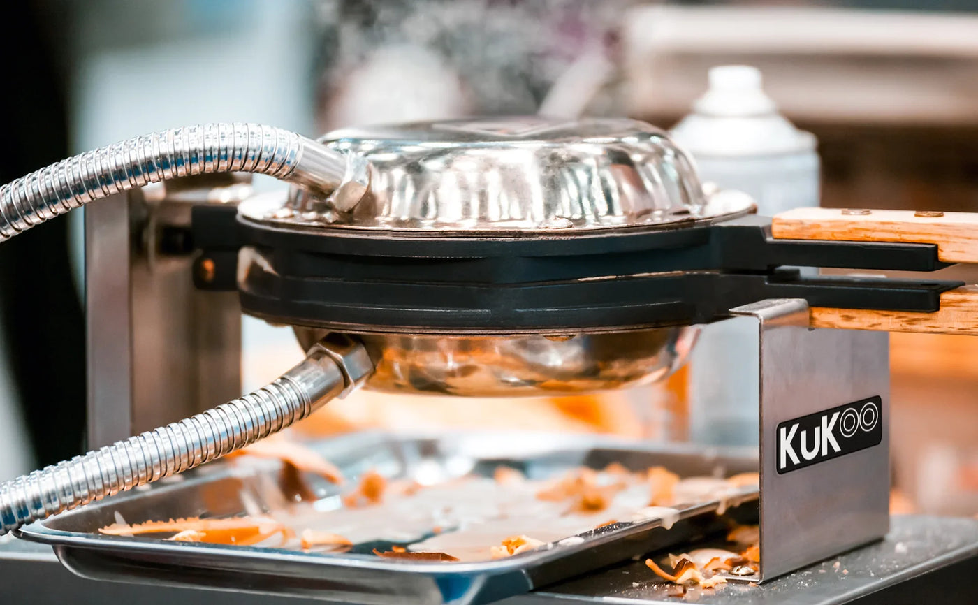 A shiny, silver waffle maker with a wooden handle, resting on a metal tray with scattered pieces of caramel and a blurred background
