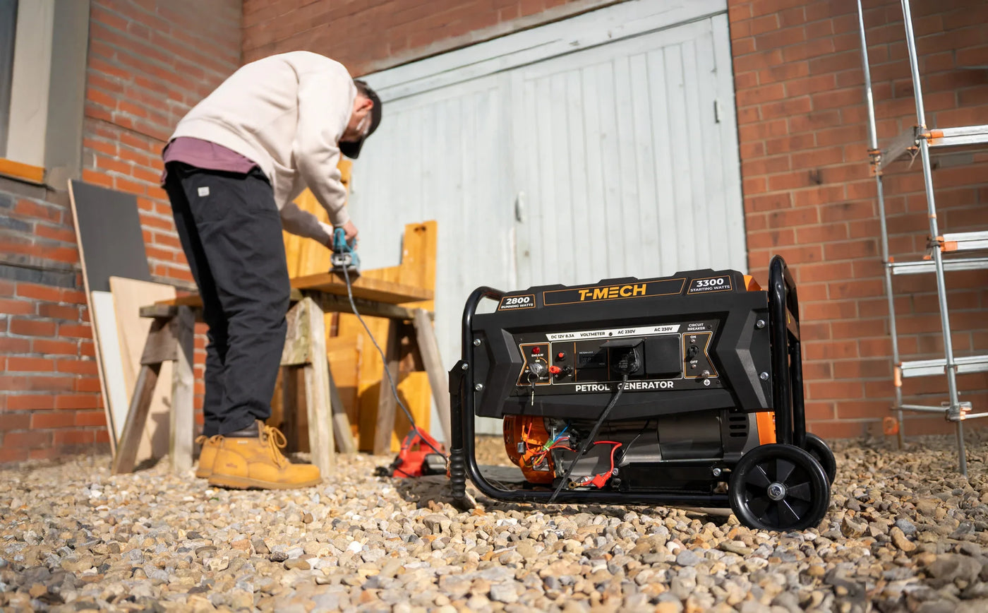 Portable T-MECH 3300 watt generator with orange accents, set on gravel, while a person in work boots uses a drill nearby