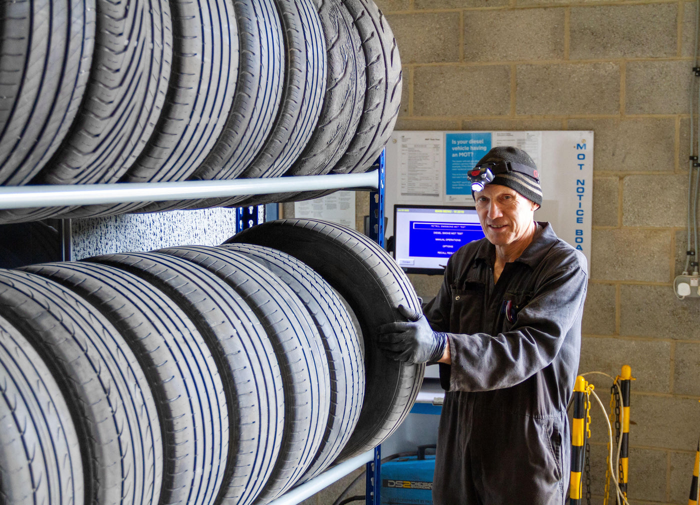 A mechanic in a black jumpsuit holds a tire while standing next to a rack of stacked tires in a garage setting