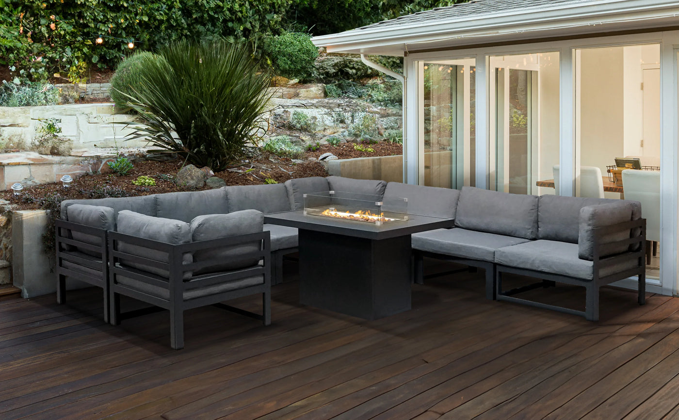Modern outdoor seating set with gray cushions, black frame, and a central fire pit, surrounded by lush greenery and wooden decking