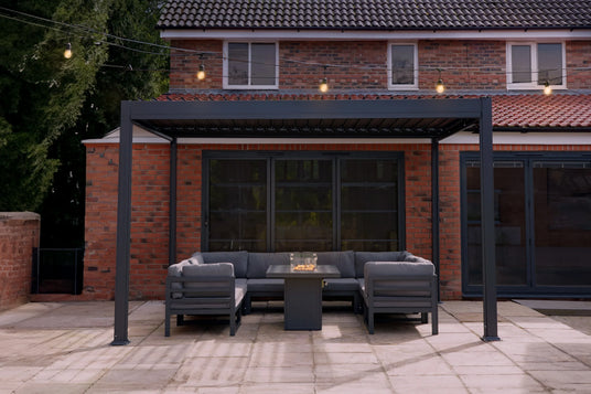 Modern outdoor seating area featuring a gray sectional sofa, fire pit table, and sleek black pergola against a brick backdrop