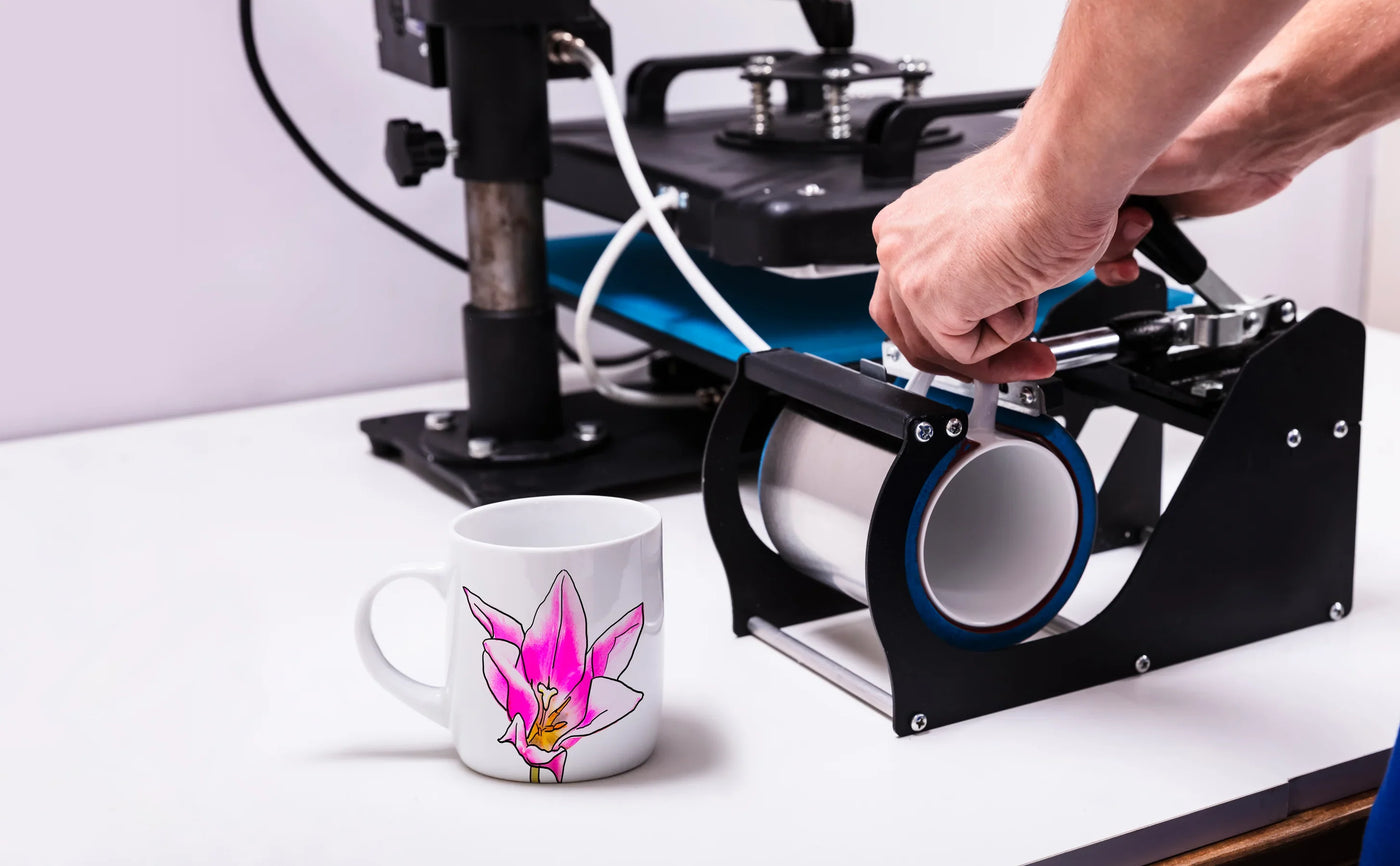 A hand operates a mug press machine, with a white mug featuring a vibrant pink orchid design on a clean workspace