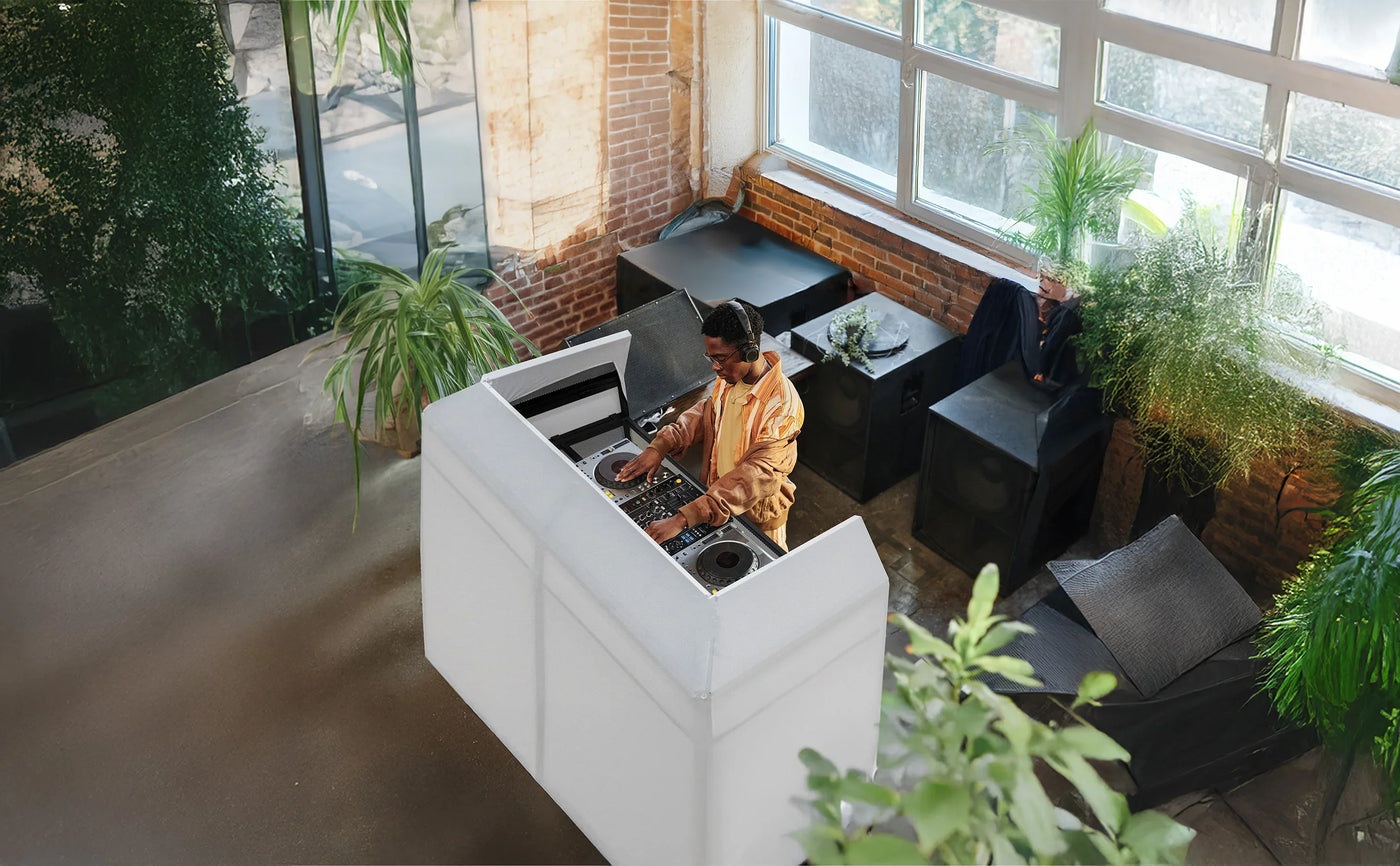 A DJ in a stylish indoor space mixes music at a sleek white booth surrounded by greenery and modern furniture