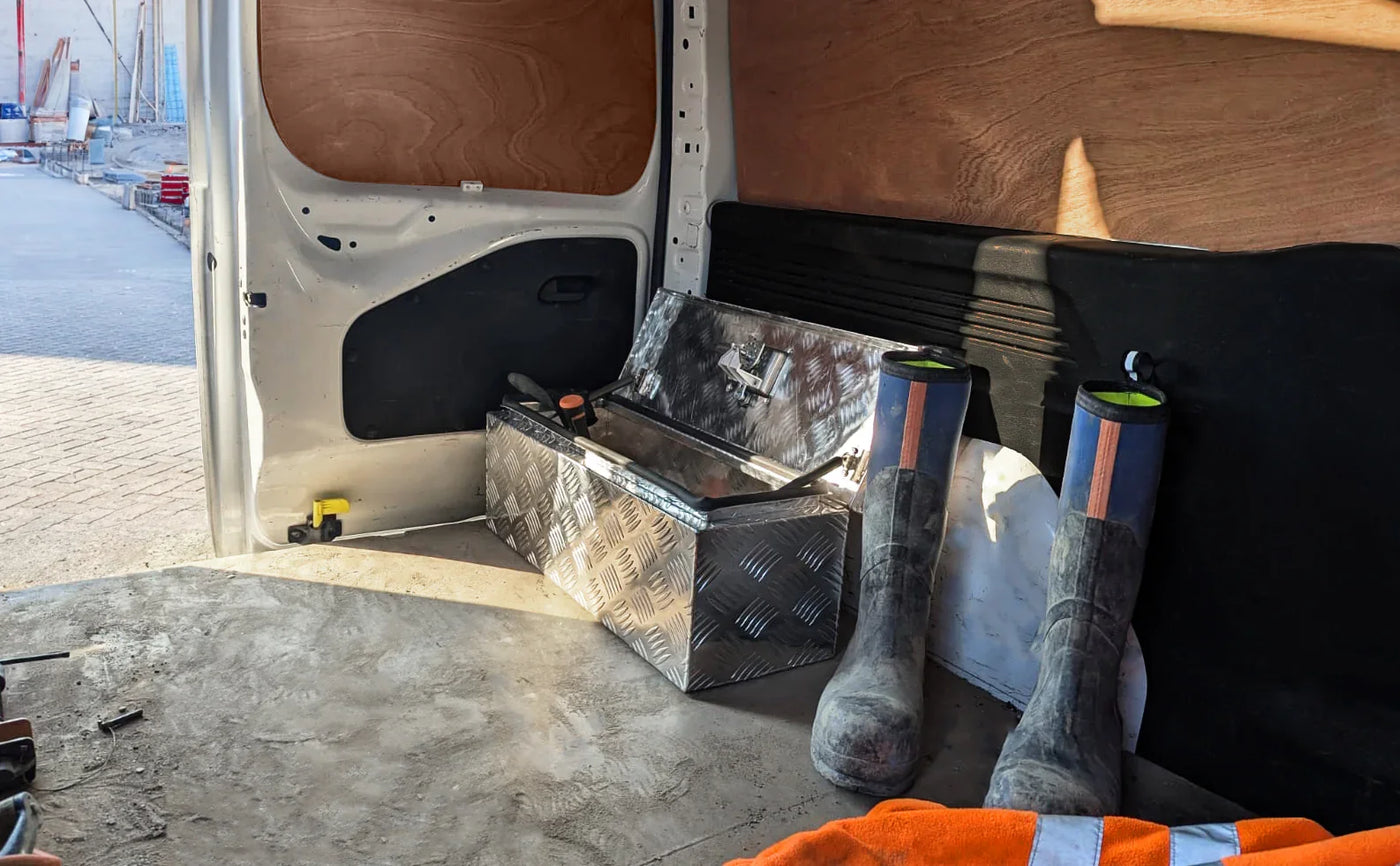 Metal toolbox with diamond plate texture sits on the floor of a van beside muddy work boots and an orange safety vest