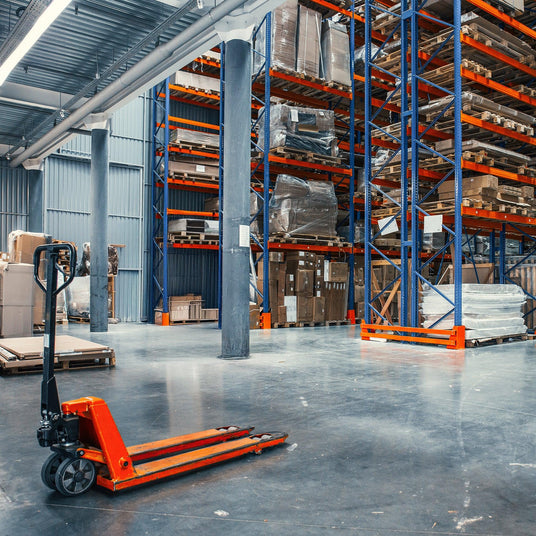 A red pallet jack on a polished concrete floor in a spacious warehouse with tall, organized shelving units filled with boxes