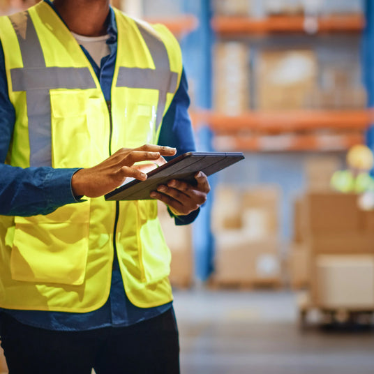 Warehouse worker in a bright yellow safety vest using a tablet, surrounded by shelves filled with cardboard boxes and pallets