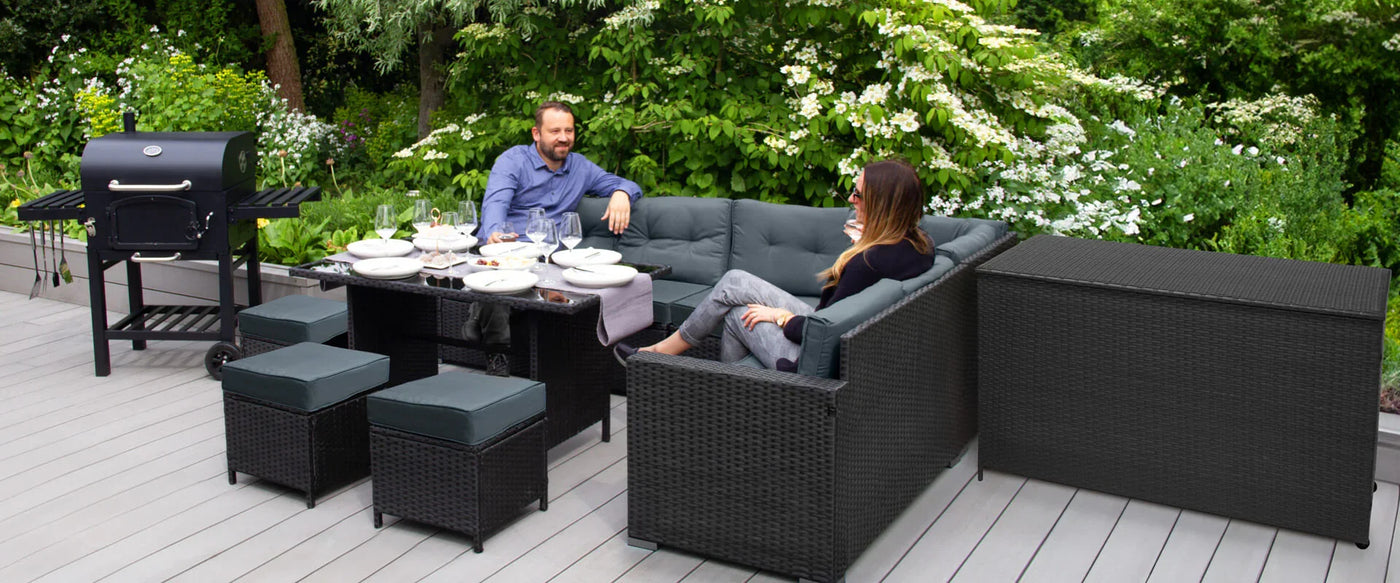 Stylish outdoor seating area with a black wicker sectional, gray cushions, and a grill, surrounded by lush greenery and flowers