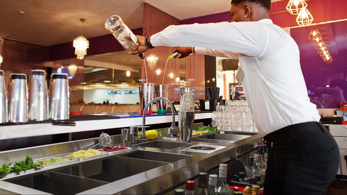 A bartender in a white long-sleeve shirt and black pants pours a drink at a bar with stainless steel sinks, metal shakers, and glassware in a modern bar setting.