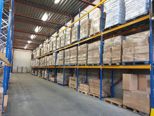 A spacious warehouse aisle lined with tall blue metal shelves stacked with neatly wrapped cardboard boxes and pallets