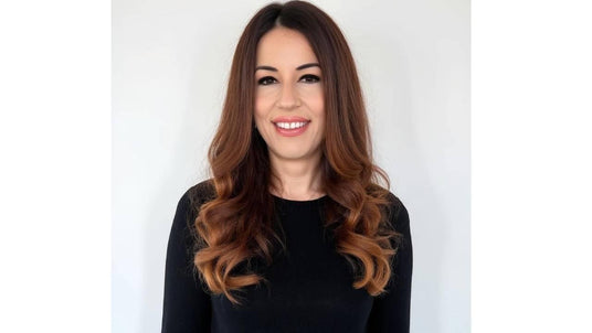 Smiling woman with long, wavy brown hair wearing a black long-sleeve top against a plain white background