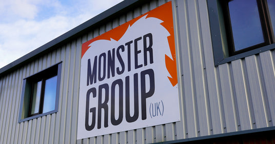 Bold orange and white sign displaying "MONSTER GROUP (UK)" on a textured gray metal building exterior with windows