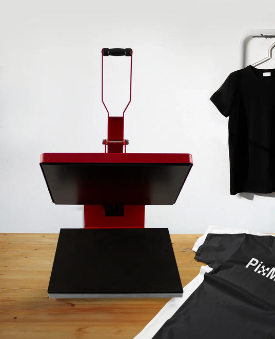 Red heat press machine on a wooden table, with a black t-shirt hanging nearby and a gray shirt laid flat, showcasing its design