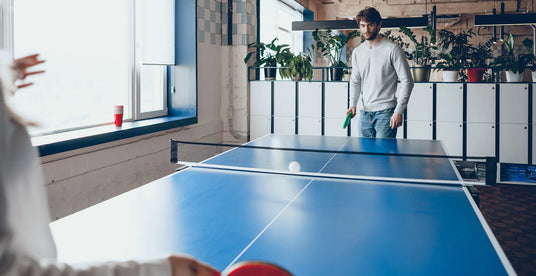 A man in a light gray sweater stands at a blue ping pong table, preparing to hit a white ball while a woman plays nearby