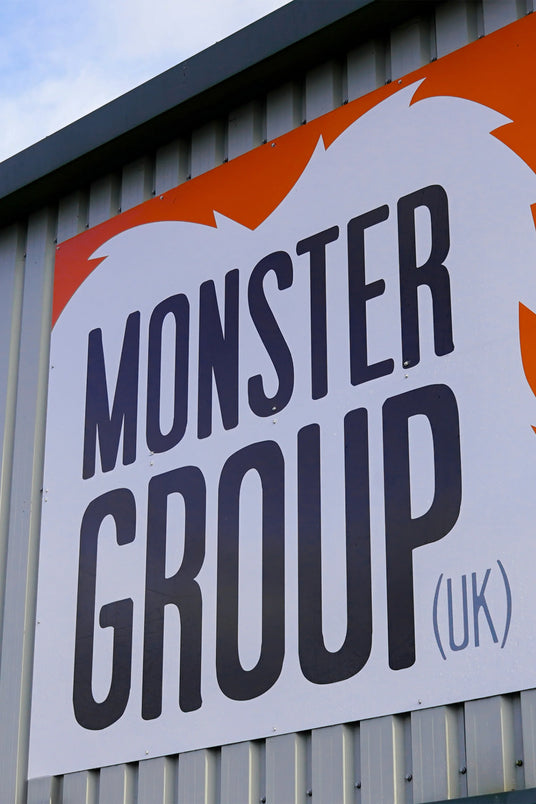 Bold, black lettering on a white sign with an orange background, displaying "MONSTER GROUP (UK)" against a corrugated metal wall