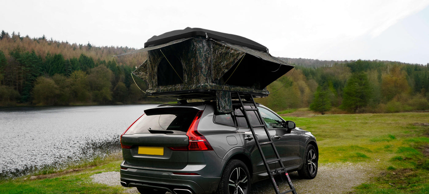 A sleek gray SUV parked by a serene lake, featuring a camouflage rooftop tent and a sturdy ladder for easy access