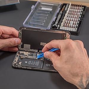 Precision screwdriver set in use, featuring a hand using a blue plastic prying tool on a smartphone's battery compartment
