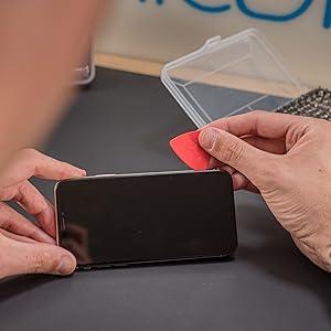 Hands using a red plastic prying tool on a smartphone screen, showcasing the ilabPRO 120 Piece Precision Screwdriver Set in action