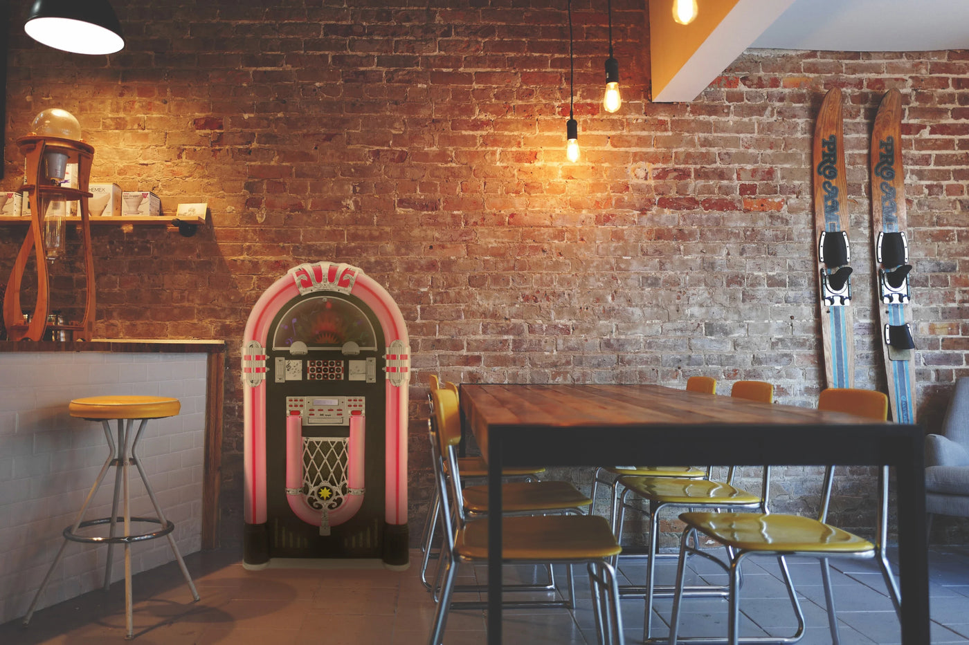 Retro jukebox with pink accents stands against a rustic brick wall, surrounded by wooden furniture and vintage ski gear