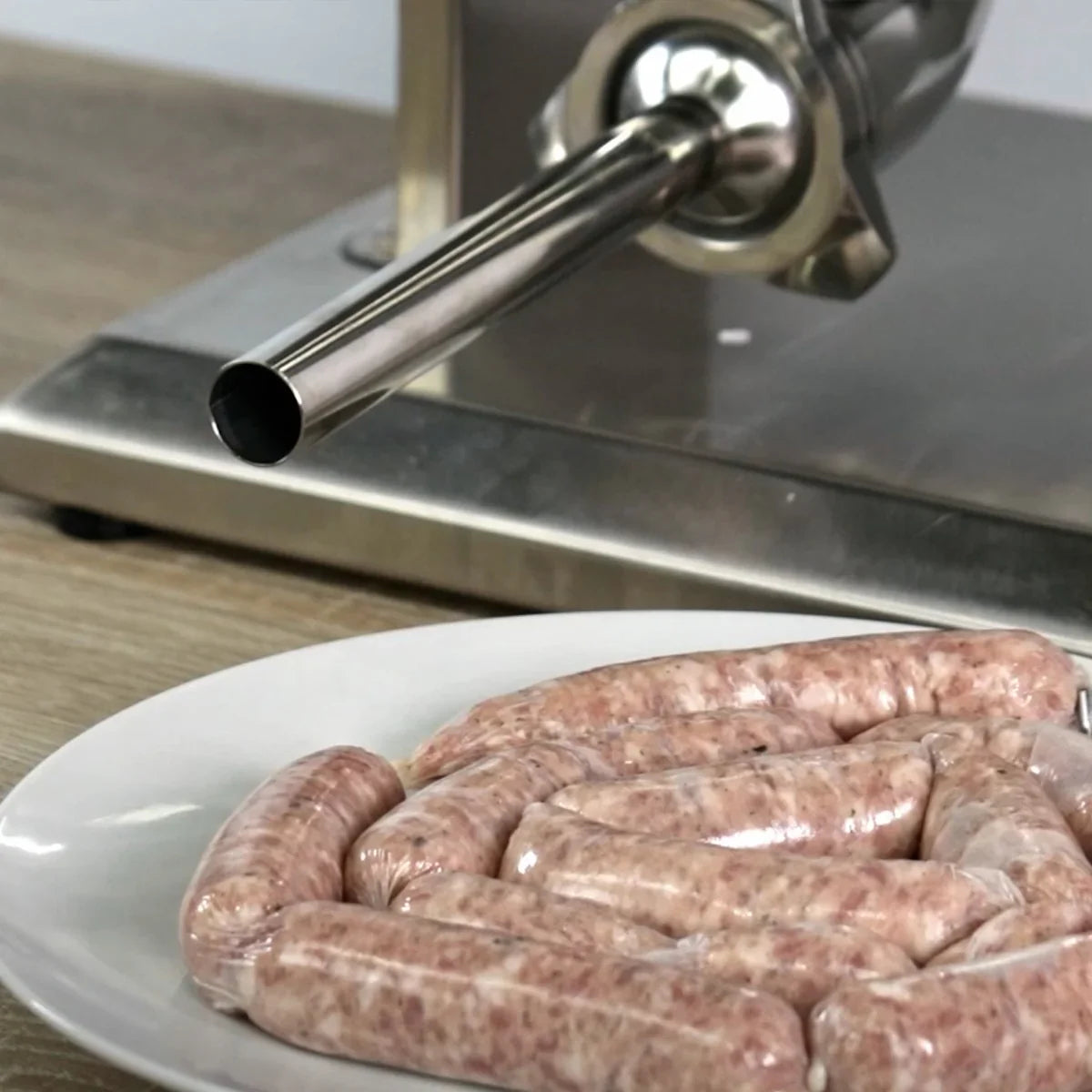 Kukoo 7 Litre Sausage Stuffer with stainless steel nozzle, positioned next to a plate of freshly made sausages on a wooden surface