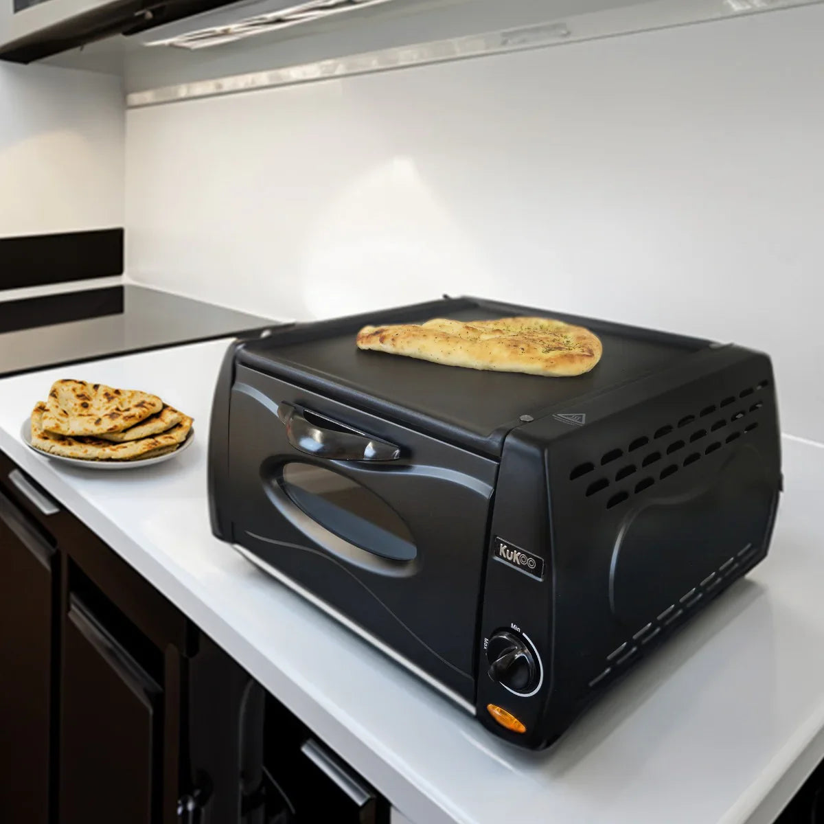 Compact black mini tandoor oven on a kitchen countertop, with naan bread cooking on top and a plate of naan beside it