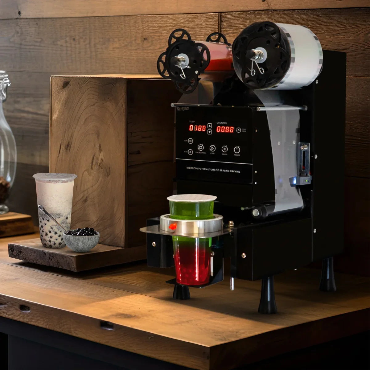 Automatic cup sealing machine in black, featuring digital display, two sealing heads, and a colorful cup with layered drinks on a wooden countertop