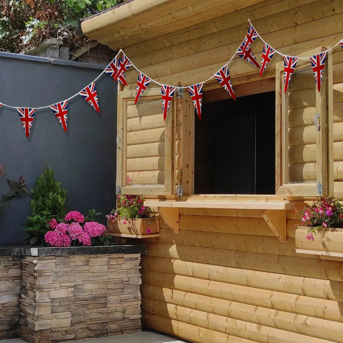 Union Jack flag bunting strung across a wooden cabin window, vibrant red, white, and blue flags complementing pink flowers below