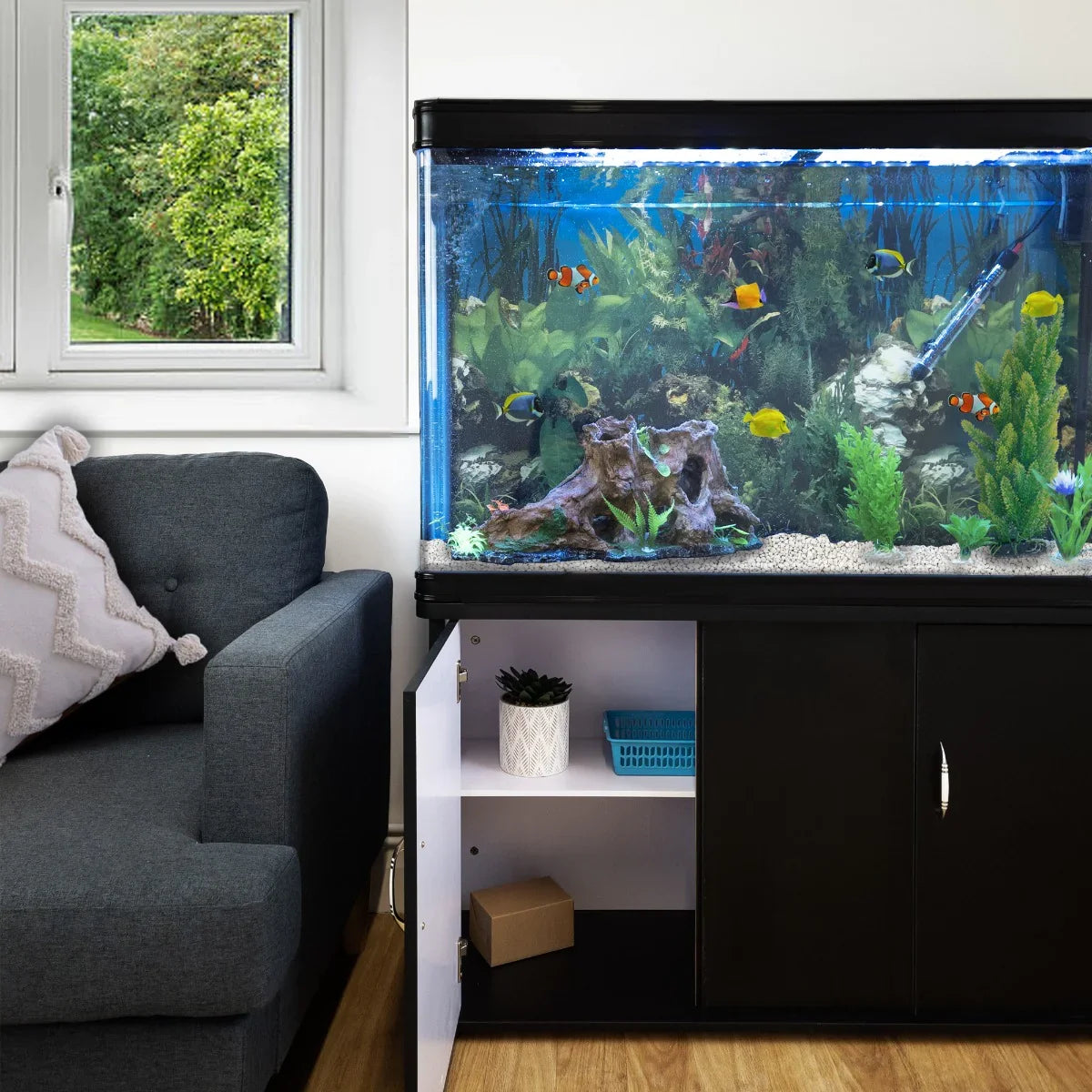 Aquarium fish tank with black cabinet, vibrant fish, white gravel, and lush plants, positioned next to a cozy sofa and window view