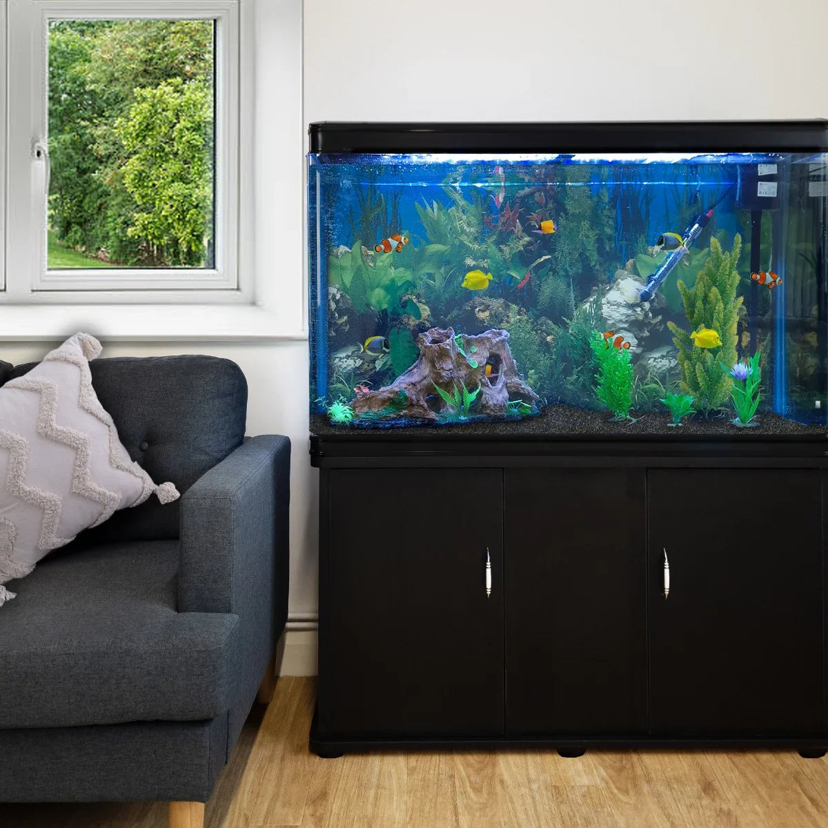 Black aquarium fish tank with vibrant plants and colorful fish, set on a sleek cabinet beside a cozy gray sofa and window