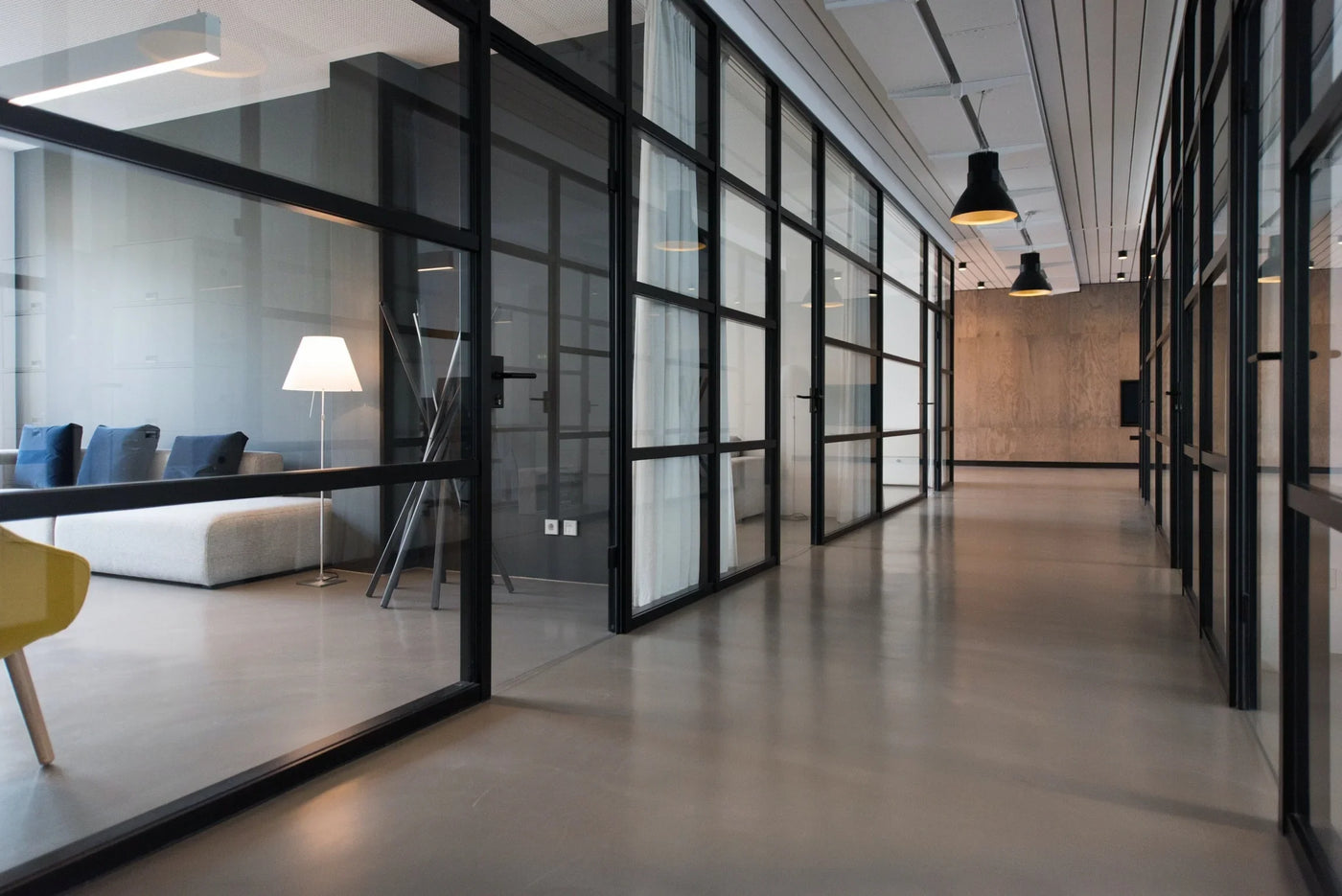 Modern office hallway featuring glass partitions, sleek concrete floors, and stylish pendant lighting, creating a bright, open atmosphere