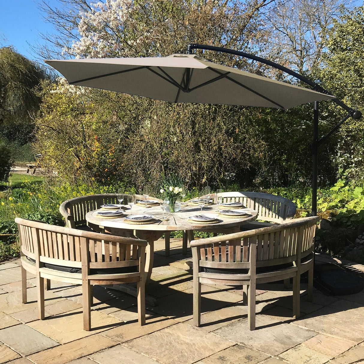 Beige cantilever parasol providing shade over a round wooden dining table set with plates and flowers, surrounded by rustic chairs