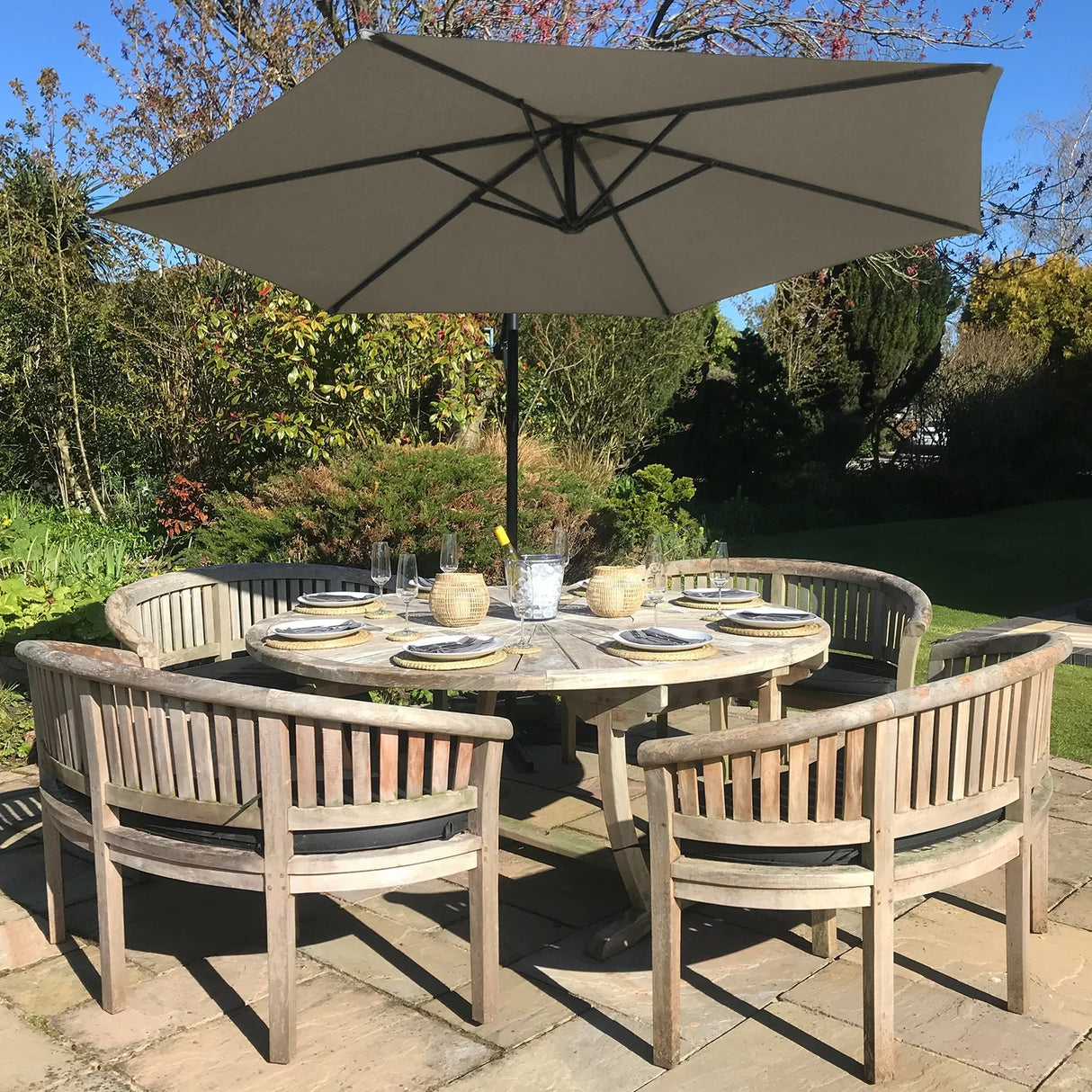 Beige cantilever parasol with a wide canopy, set above a rustic wooden dining table surrounded by curved wooden chairs in a garden setting